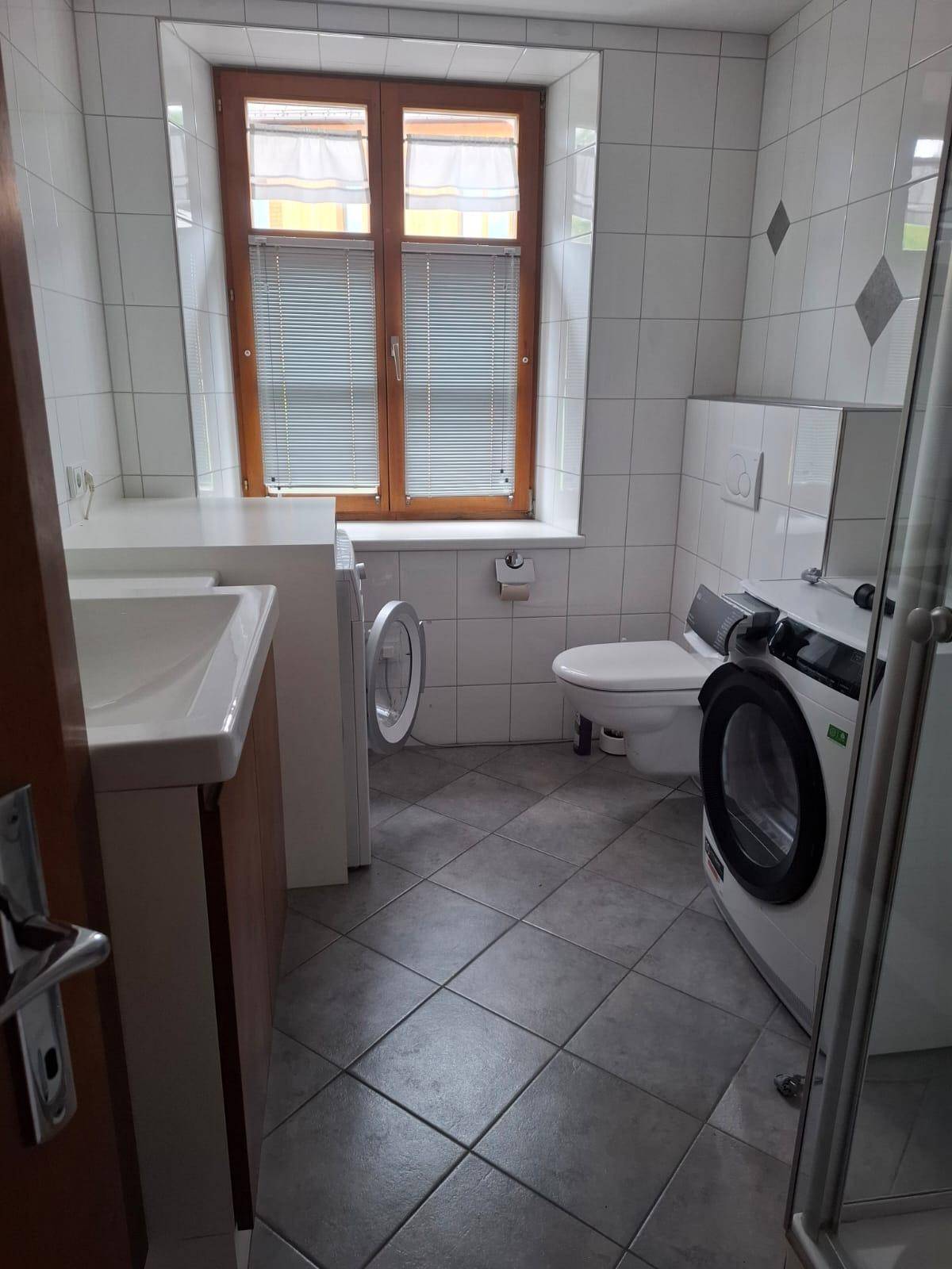 Funktionales Badezimmer mit weißen Fliesen, Fenster, Toilette und Platz für eine Waschmaschine.