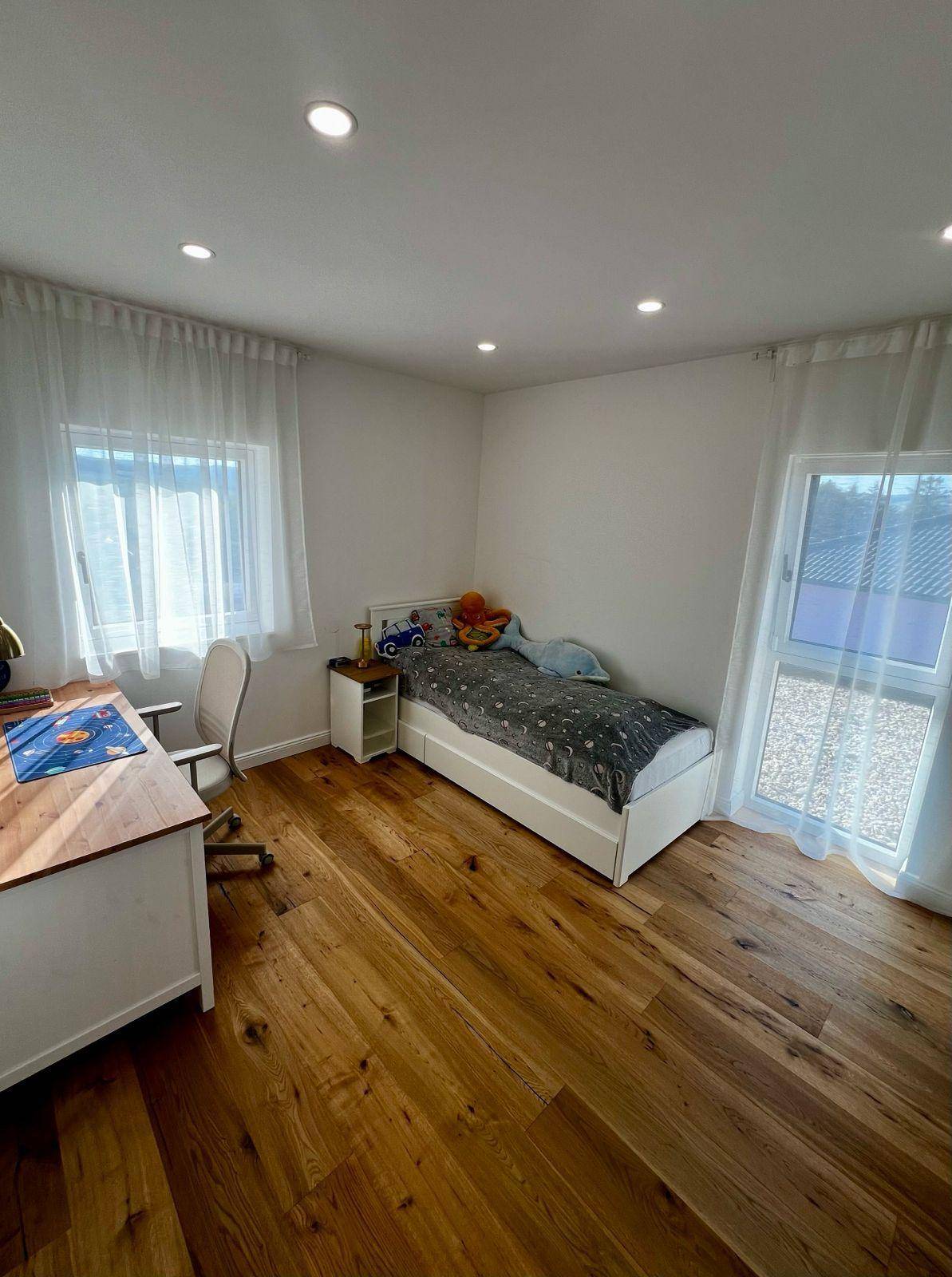 Helles Kinderzimmer mit Schreibtisch, Holzboden und Zugang zum Balkon.