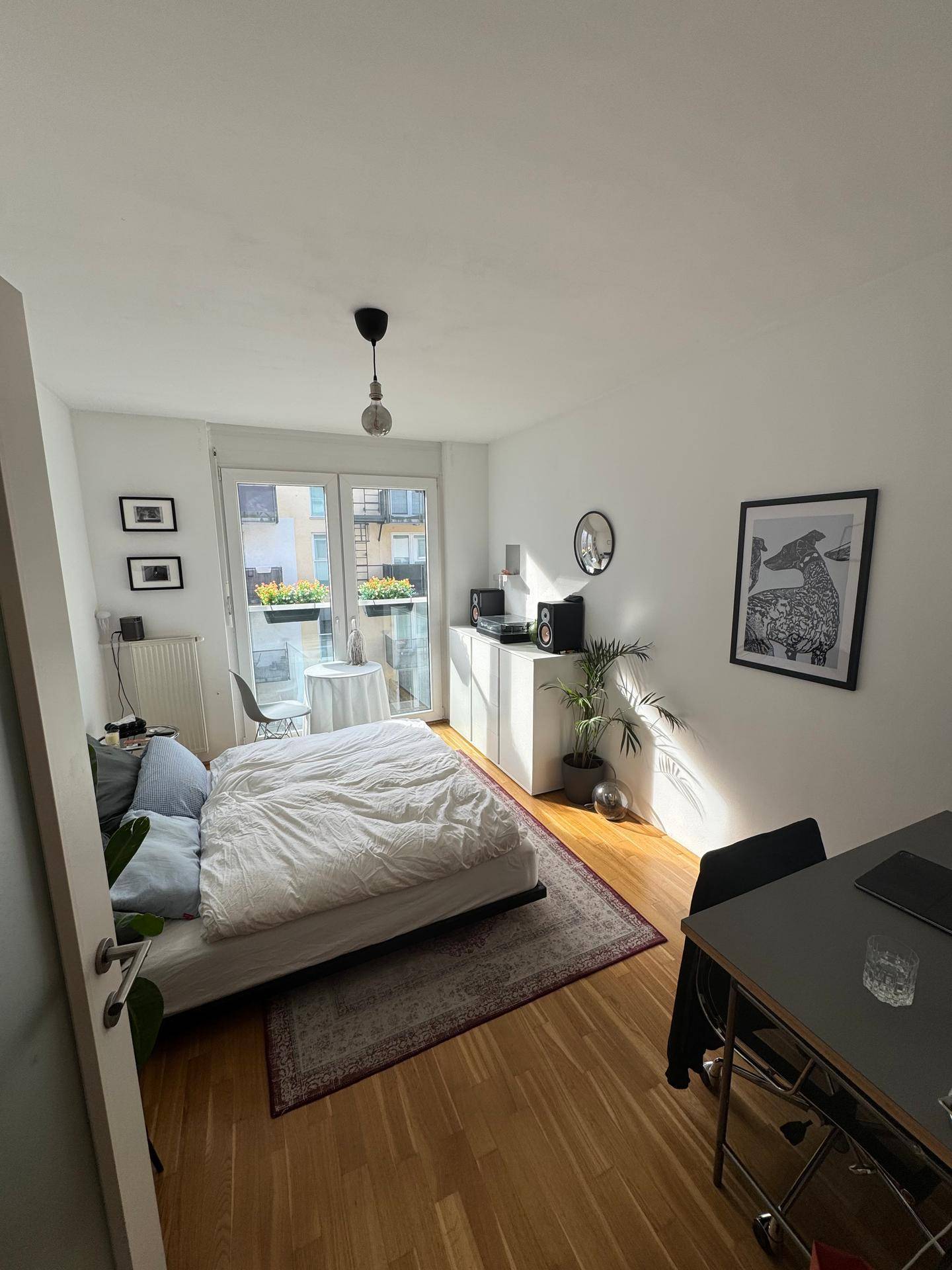 Helles Schlafzimmer mit Bett, Schreibtisch und Balkonzugang, eingerichtet mit Parkettboden.