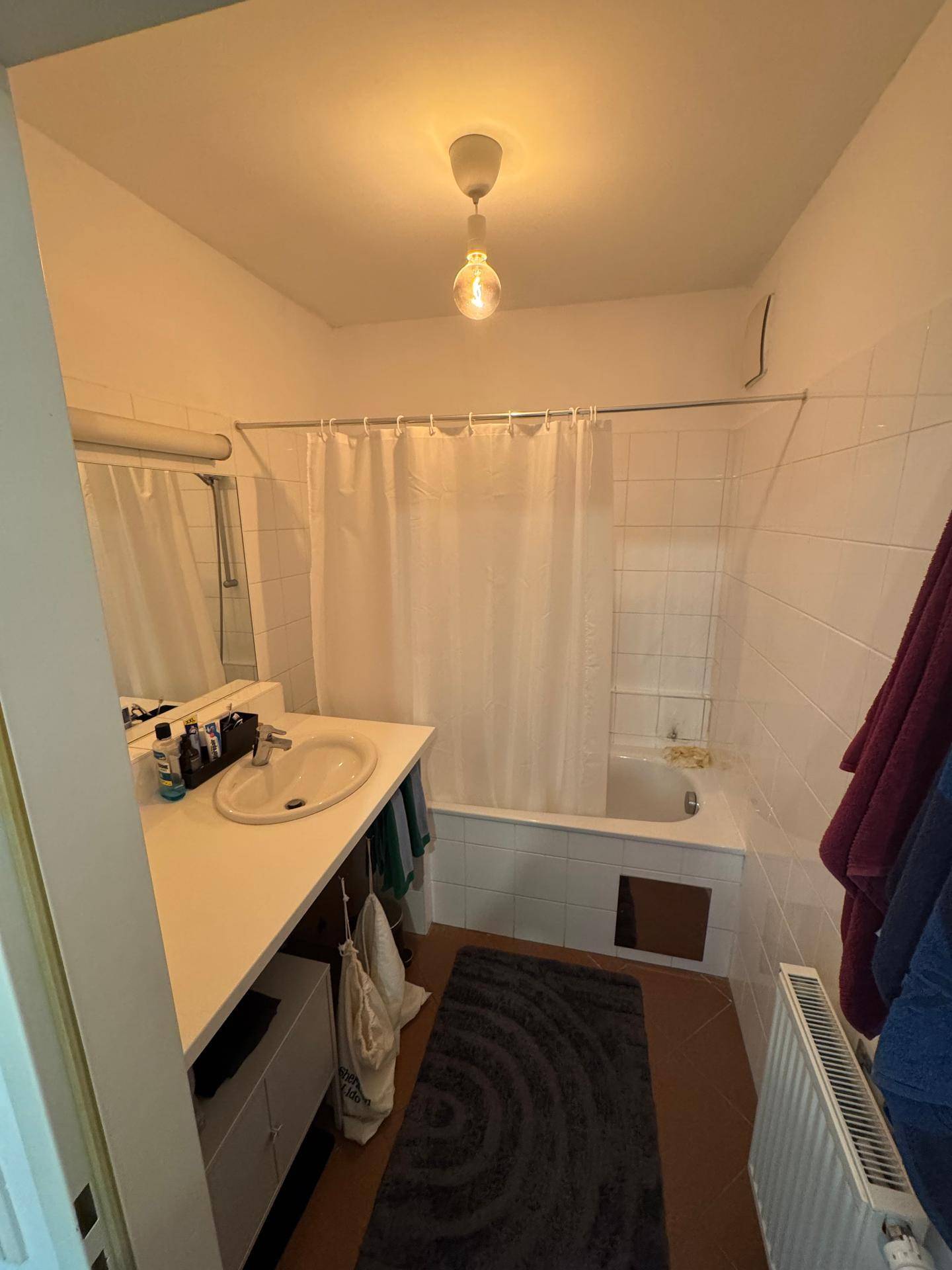 Badezimmer mit Badewanne, Waschbecken und Spiegel, ausgestattet mit weißen Fliesen und braunem Boden.