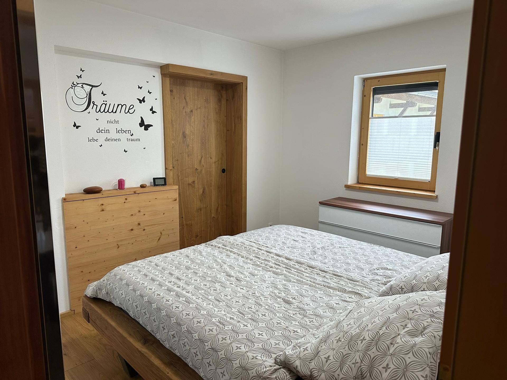 Helles Schlafzimmer mit Holzbett, Kommode und Wanddekoration in ruhiger und einladender Atmosphäre.