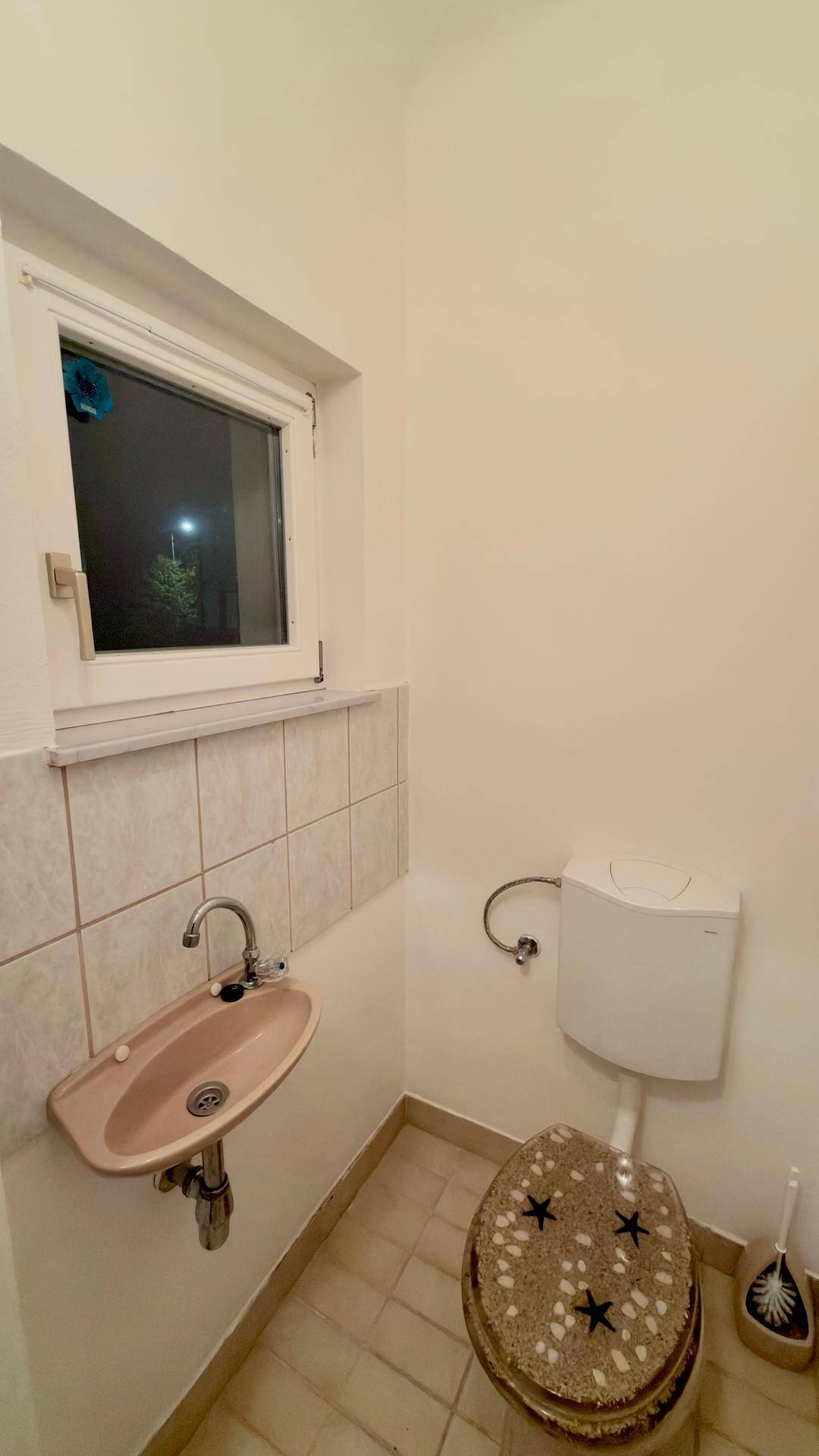 Kleines WC mit Fenster, Waschbecken und einer Toilette mit gemustertem Deckel.