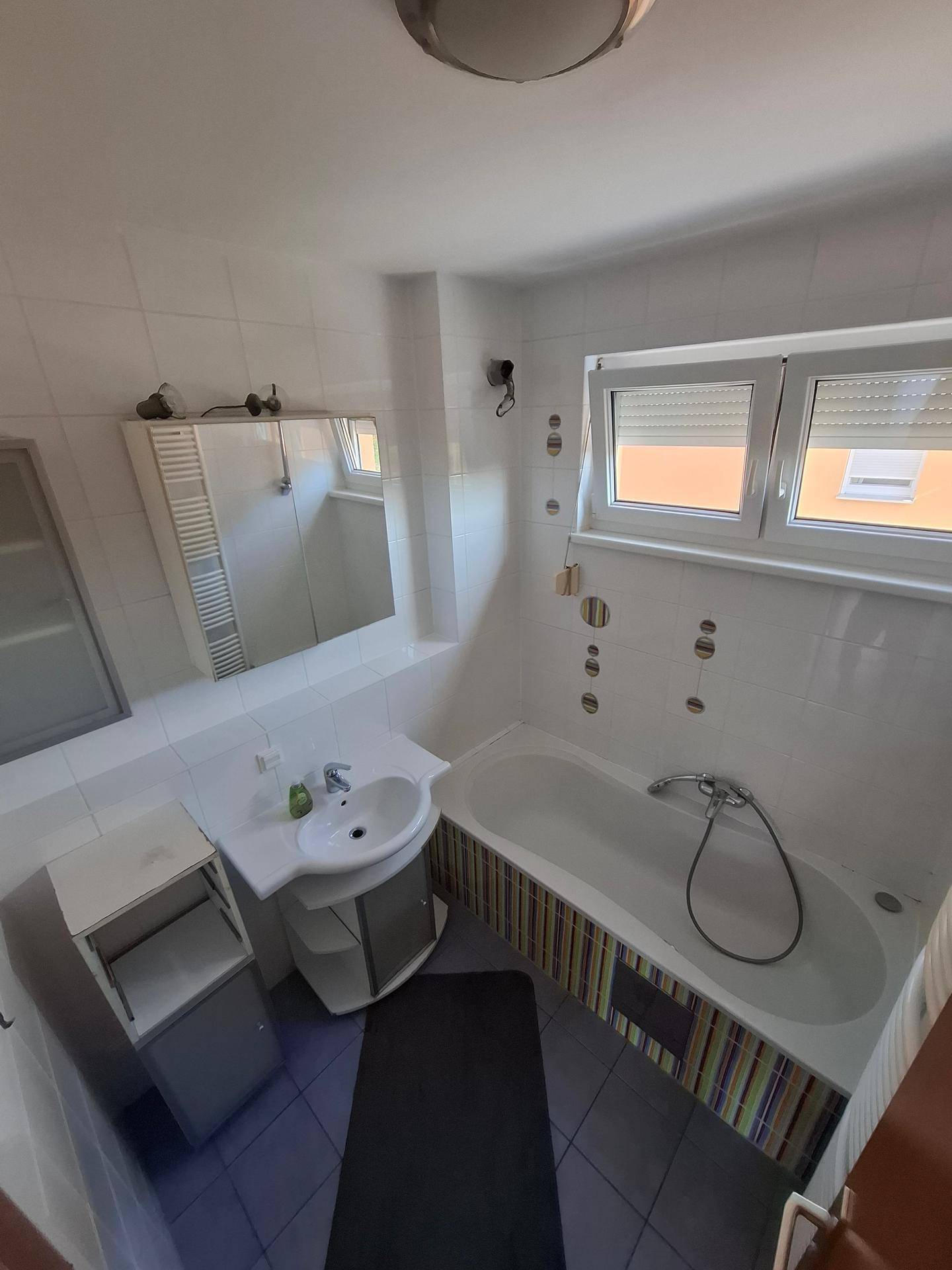 Badezimmer mit Badewanne, Einzelwaschbecken, Spiegelschrank und zwei Fenstern für Tageslicht.