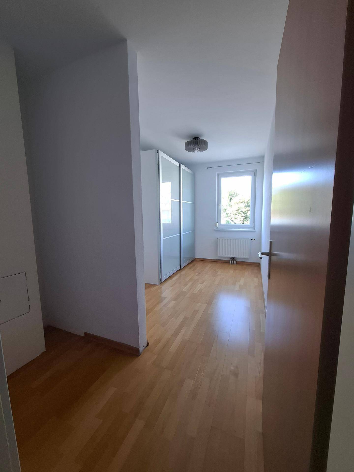 Ansicht des Schlafzimmers mit Fenster, Heizkörper und Holzboden, das viel Tageslicht bietet.