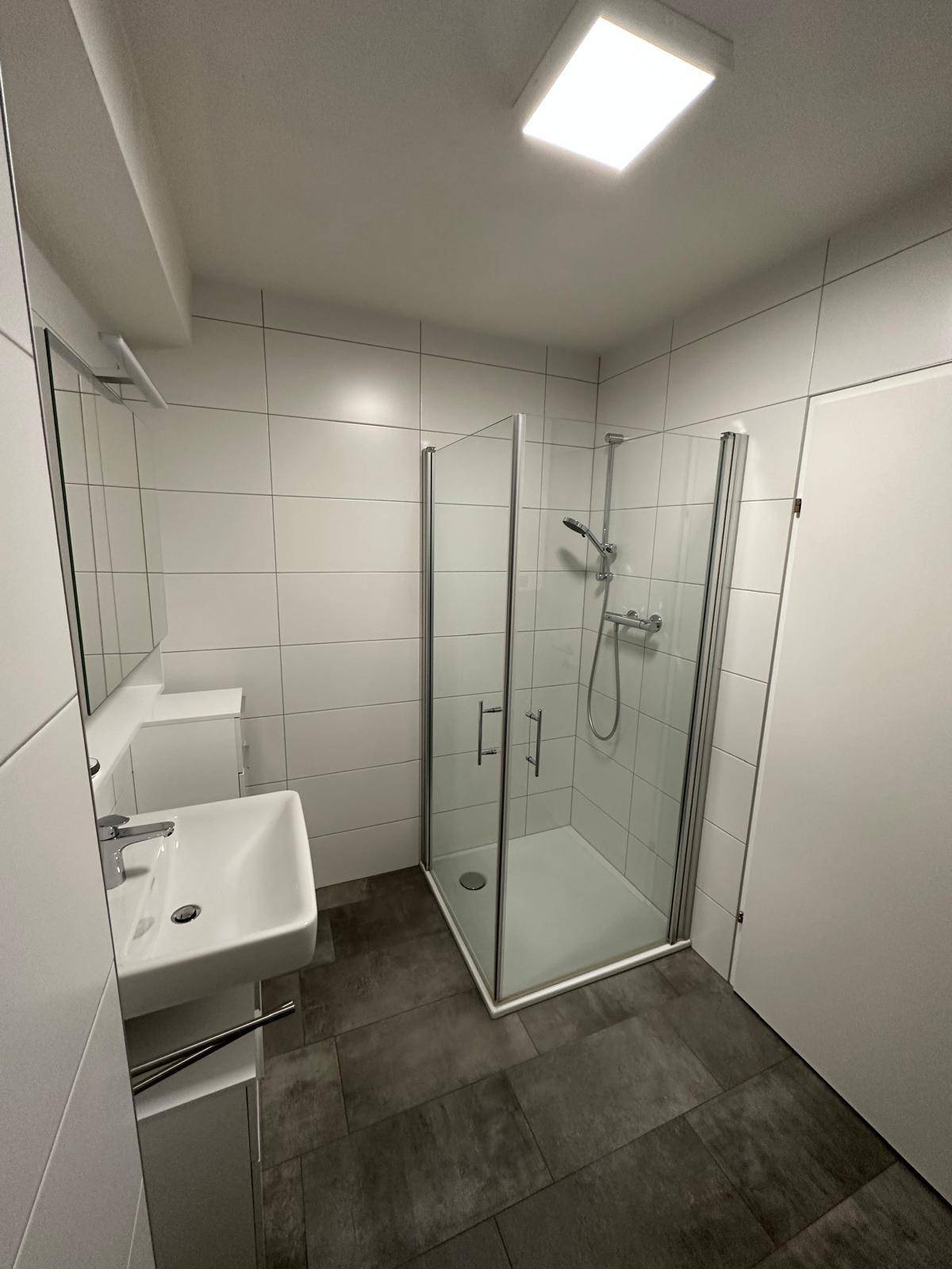Helles, modernes Badezimmer mit Duschkabine, weißen Wandfliesen und dunklem Fliesenboden.