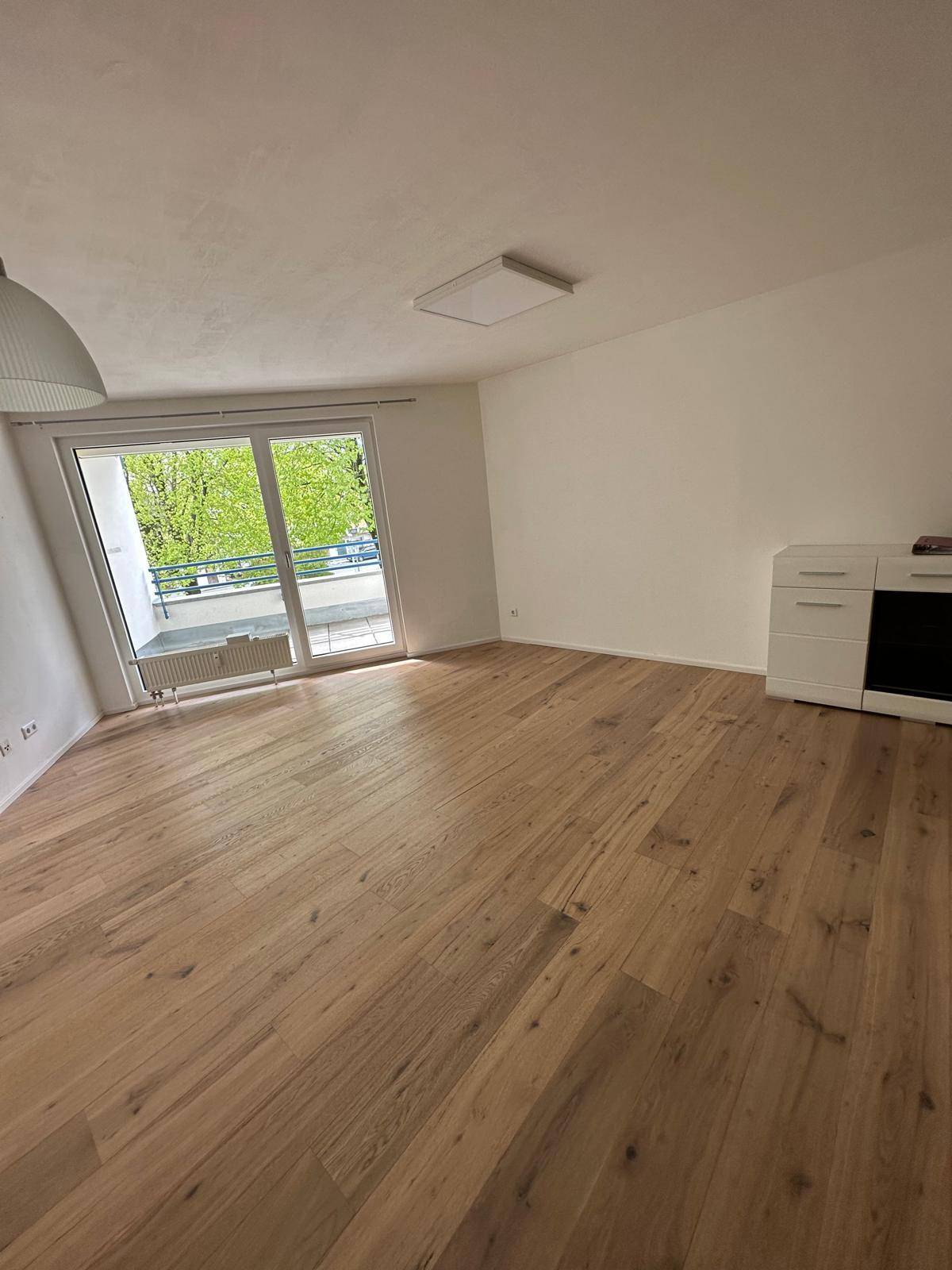 Heller Wohnbereich mit Holzboden und großem Fenster, das viel Tageslicht hereinlässt und Zugang zum Balkon bietet.