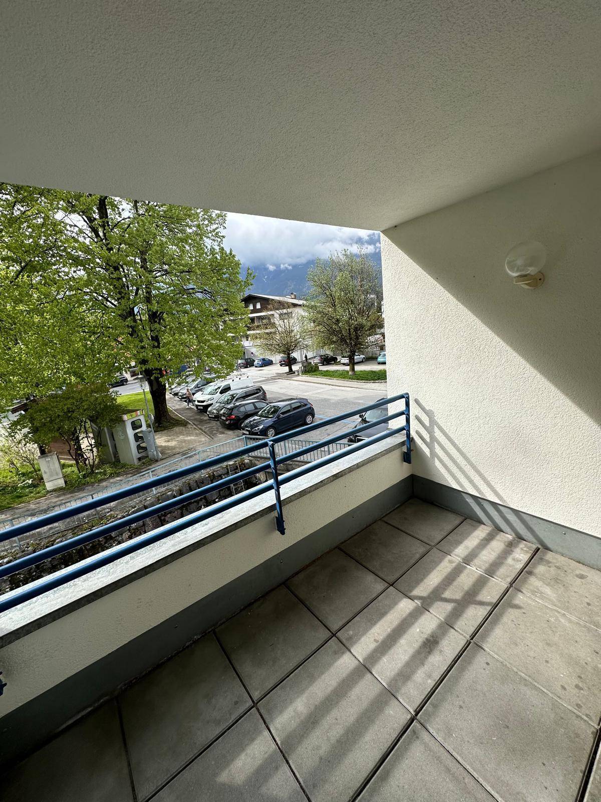 Sonniger Balkon mit Fliesenboden und blauem Geländer, bietet Blick auf Bäume und Berge.