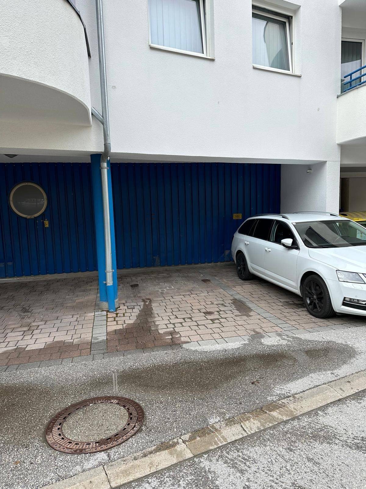 Außenansicht des Gebäudes mit blauer Garagentür und gepflastertem Bereich für Fahrzeuge.