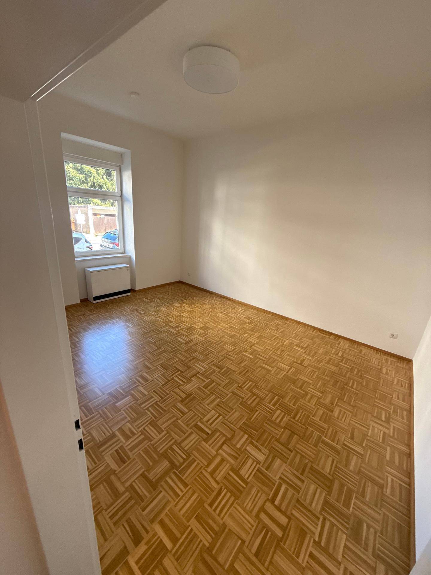 Helles Zimmer mit Parkettboden und einem Fenster, das viel Tageslicht hereinlässt.