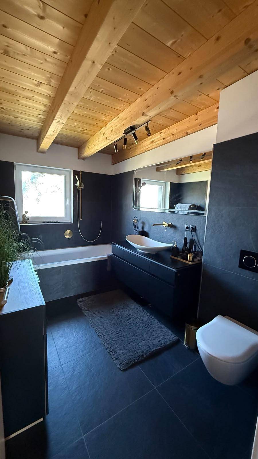 Modernes Badezimmer mit dunklen Fliesen, Badewanne, Einzelwaschbecken und Holzbalkendecke.