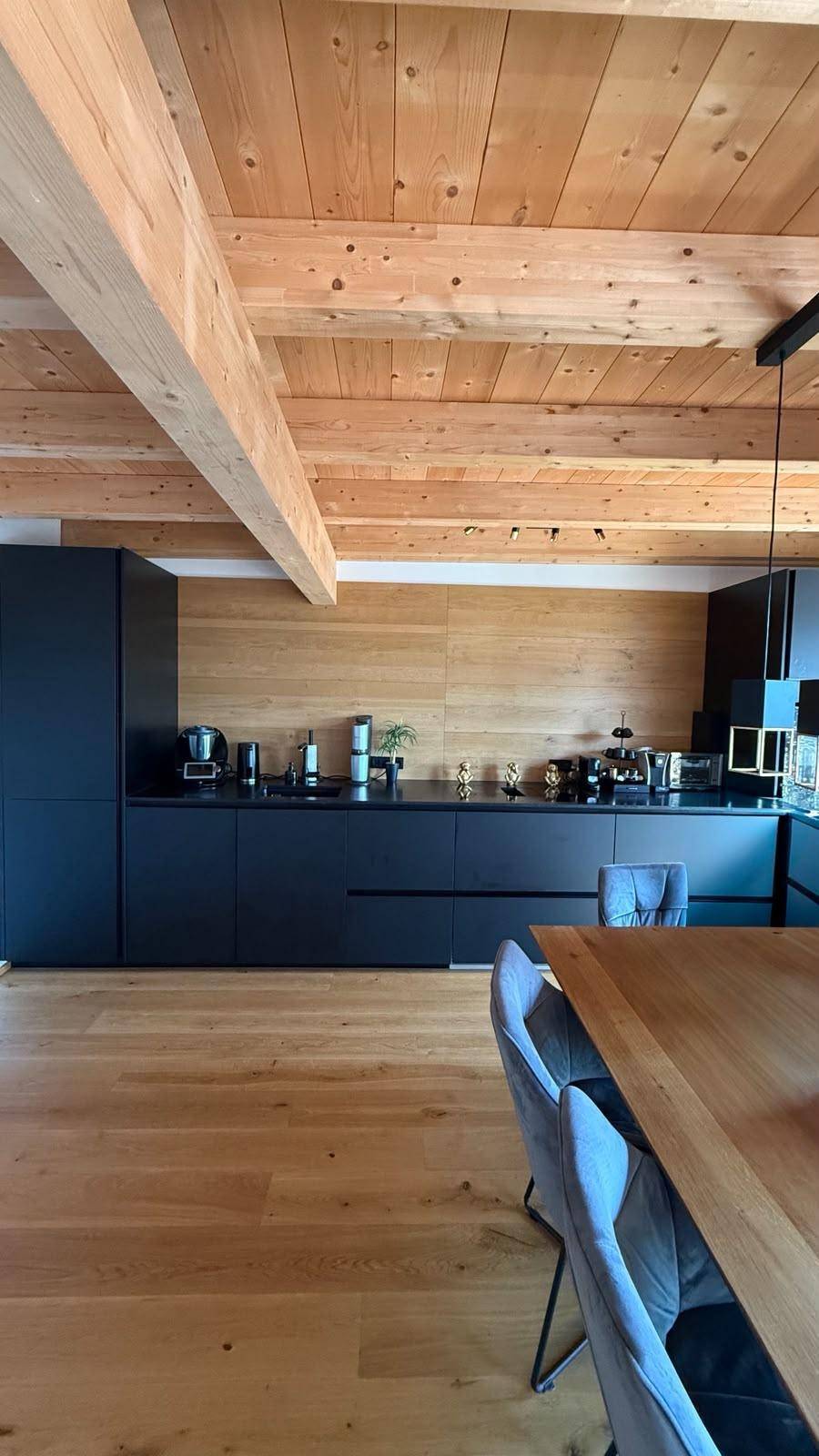 Geräumige moderne Küche mit schwarzen Fronten, Holzwand und sichtbaren Deckenbalken.
