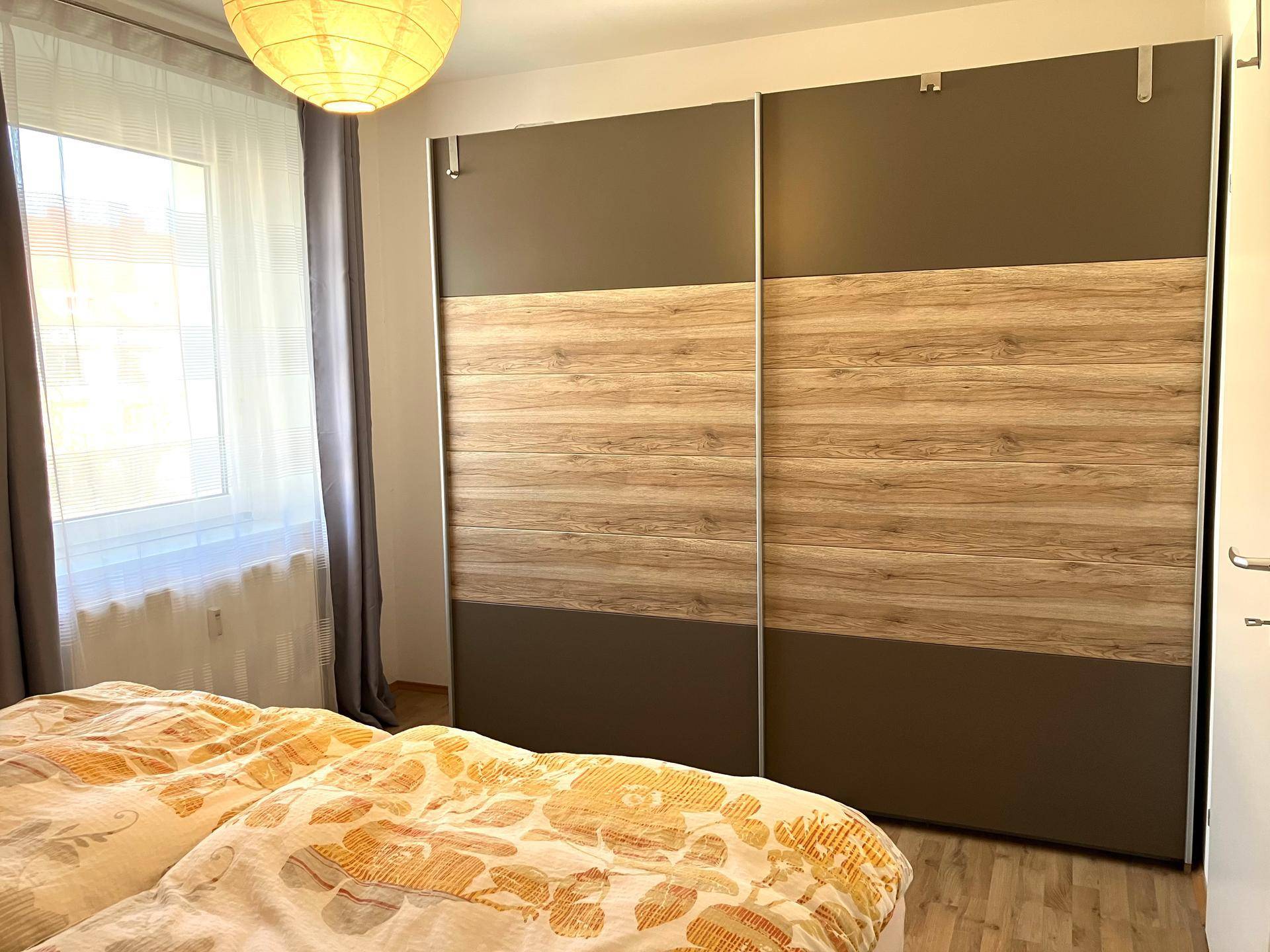Schlafzimmer mit großem Schiebeschrank in Holzoptik und Fenster mit Vorhängen, das Tageslicht hereinlässt.