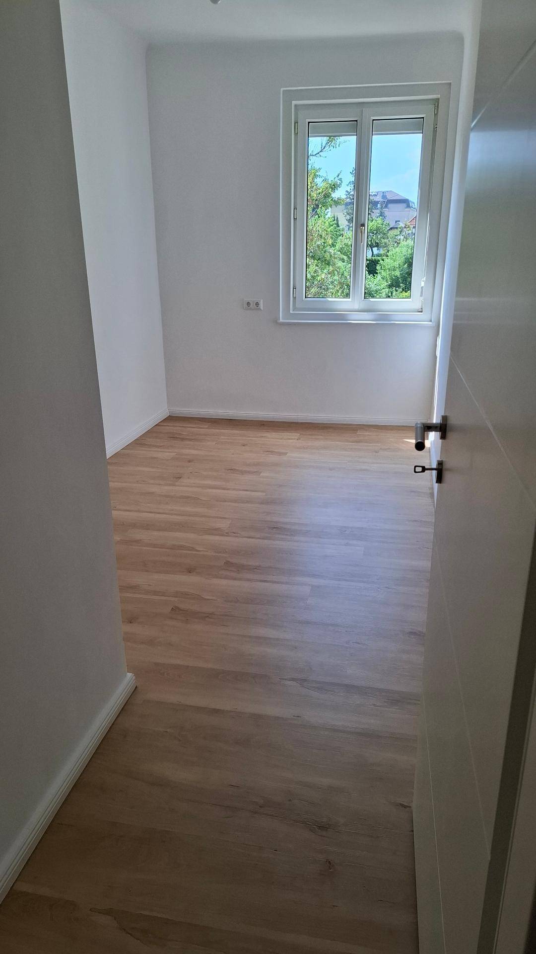 Helles, leeres Zimmer mit Holzboden und großem Fenster, bietet viel Tageslicht.