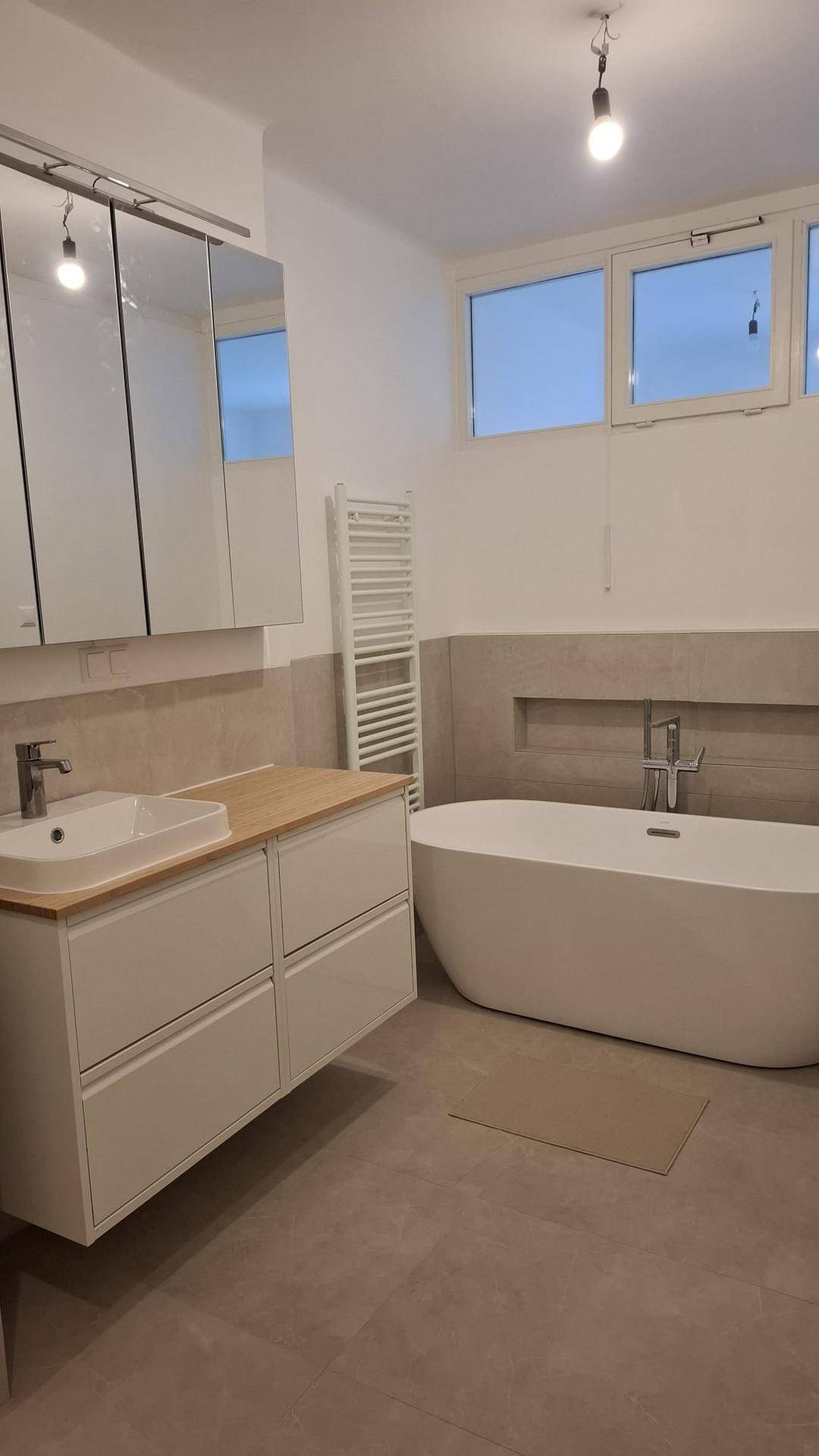Helles Badezimmer mit freistehender Badewanne, Einzelwaschbecken und Fenster, ausgestattet mit einem Handtuchheizkörper.
