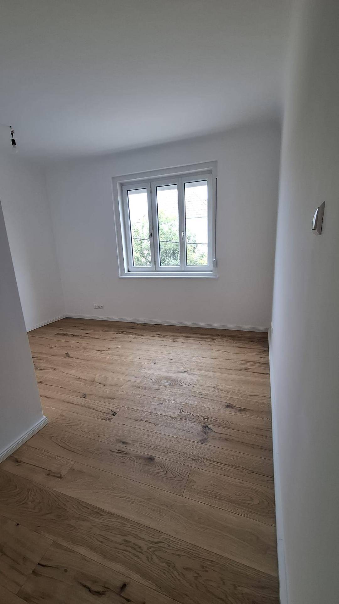 Geräumiges, helles Zimmer mit Holzboden und Fenster, bereit zur individuellen Gestaltung.