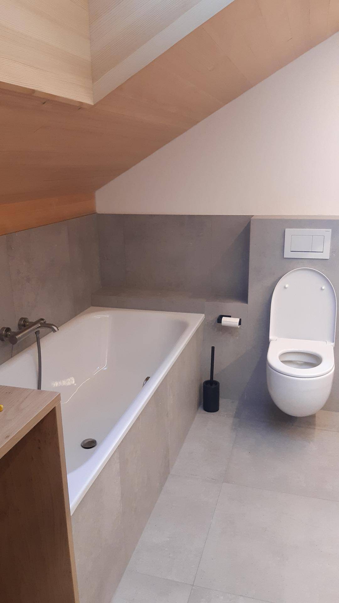 Modernes Badezimmer mit Badewanne, Wand-WC und grauen Fliesen unter einer Dachschräge.