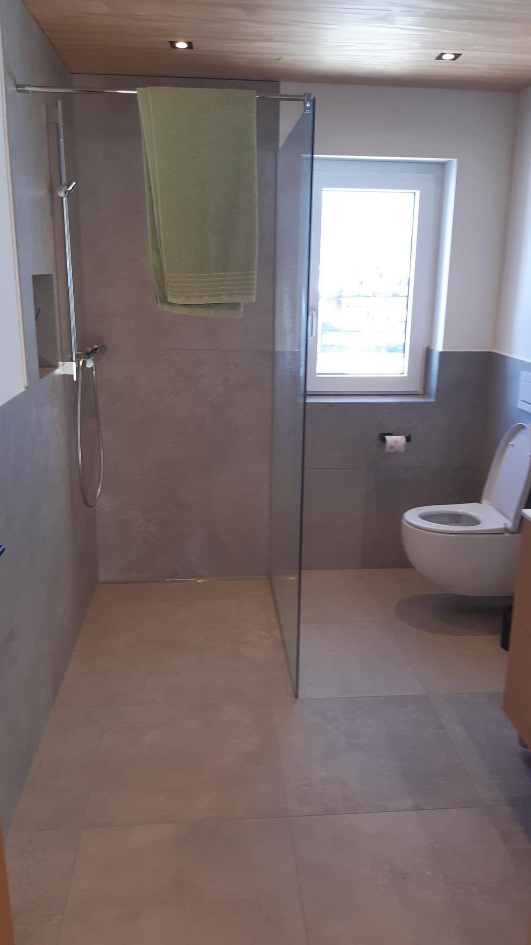 Helles Badezimmer mit bodengleicher Dusche, Wand-WC, Fenster und modernen grauen Fliesen.