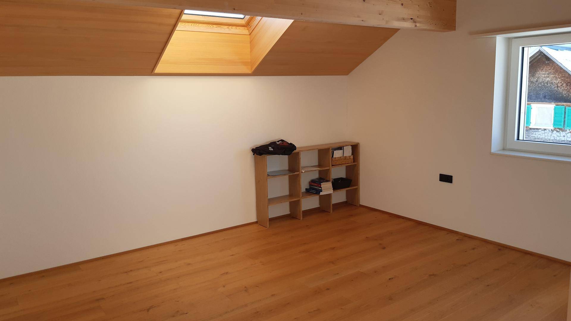 Heller Raum im Dachgeschoss mit Holzboden, einem Regal, Fenster und schräger Decke.