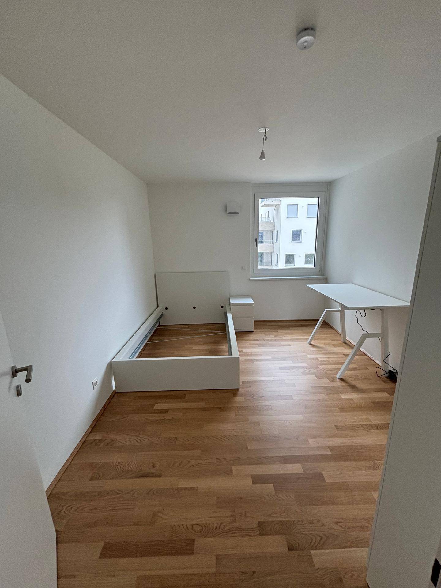Lichtdurchflutetes Schlafzimmer mit Parkettboden, Fenster und modernem Schreibtisch, ideal für Wohnen und Arbeiten.