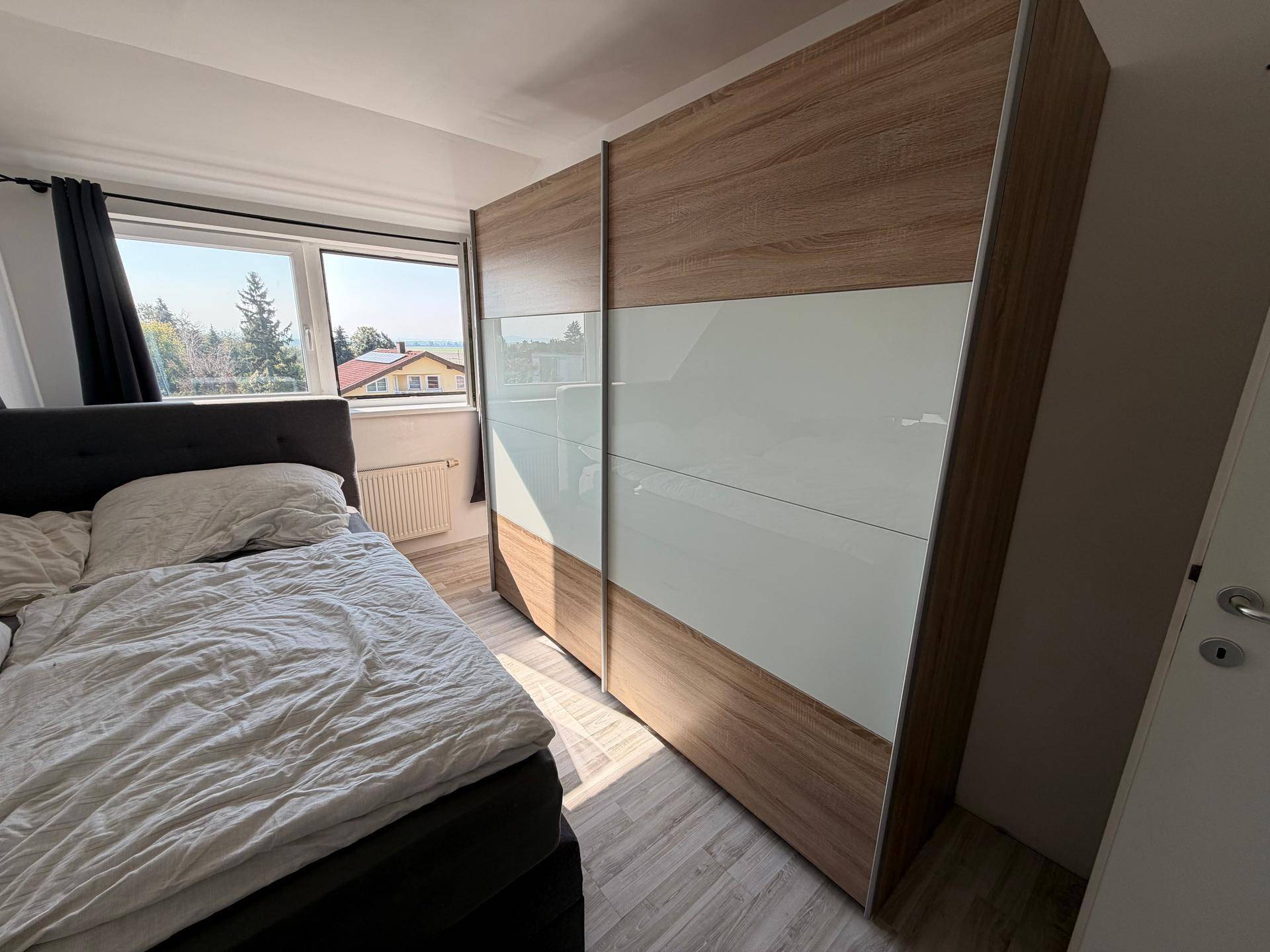 Schlafzimmer mit großem Schiebetürenschrank und Fenster, das viel Tageslicht hereinlässt und eine grüne Aussicht bietet.