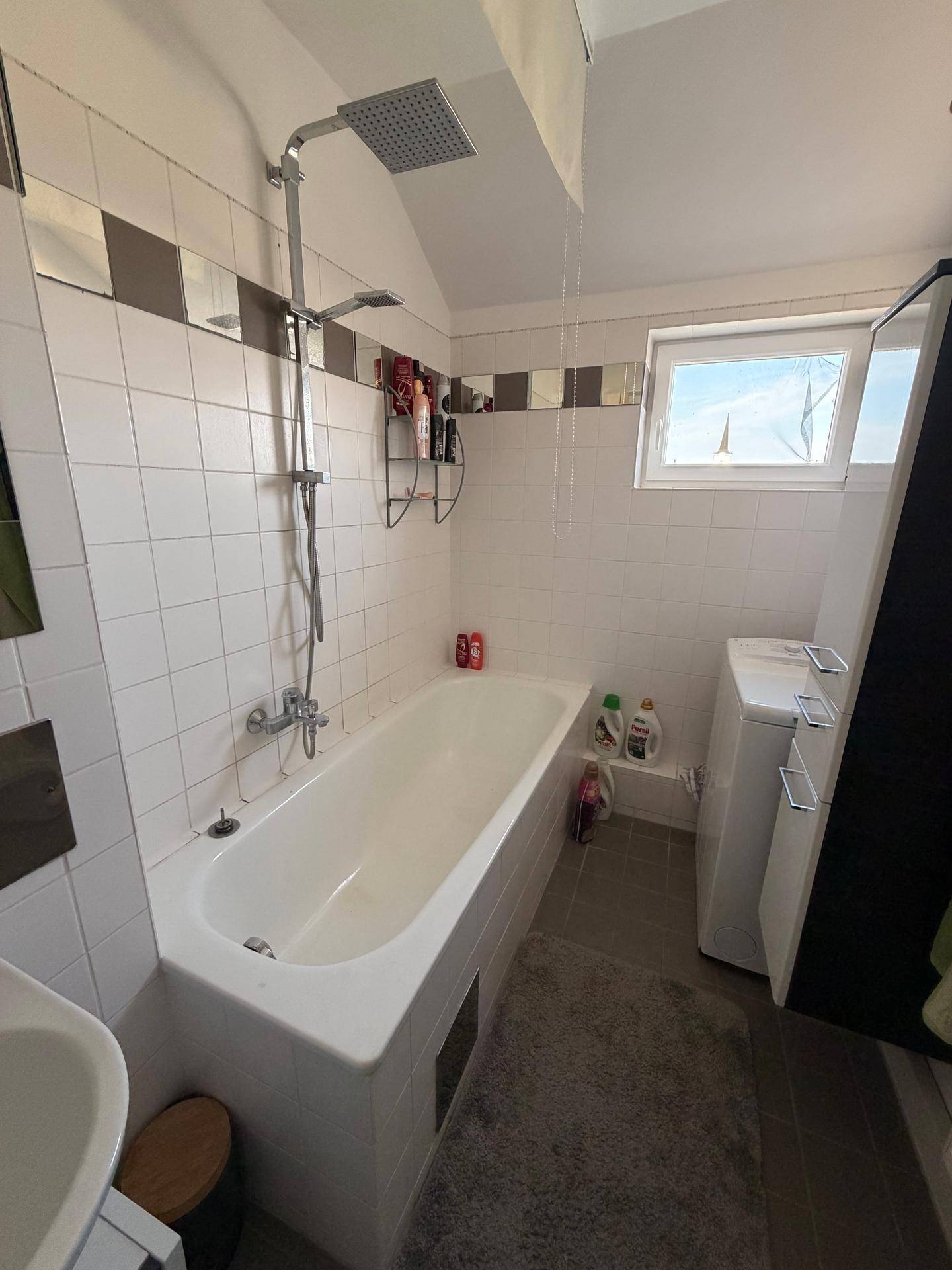 Modernes Badezimmer mit Badewanne, Regendusche und Fenster, das Tageslicht hereinlässt und eine Waschmaschine zeigt.