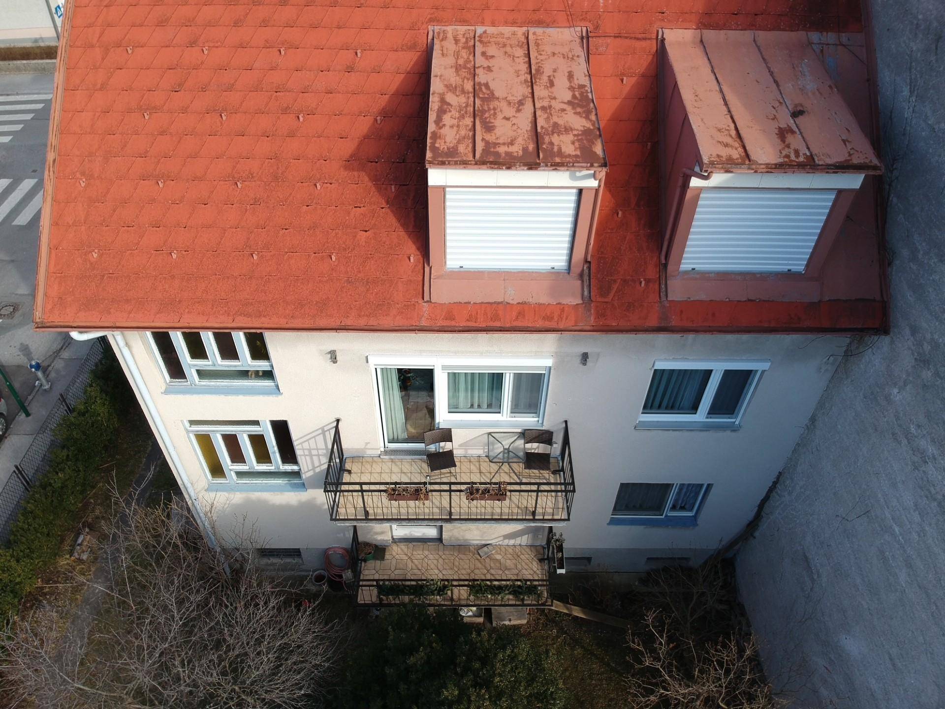 Ansicht des Hauses von oben mit Balkon, rotem Ziegeldach und Dachgauben, umgeben von Bäumen.