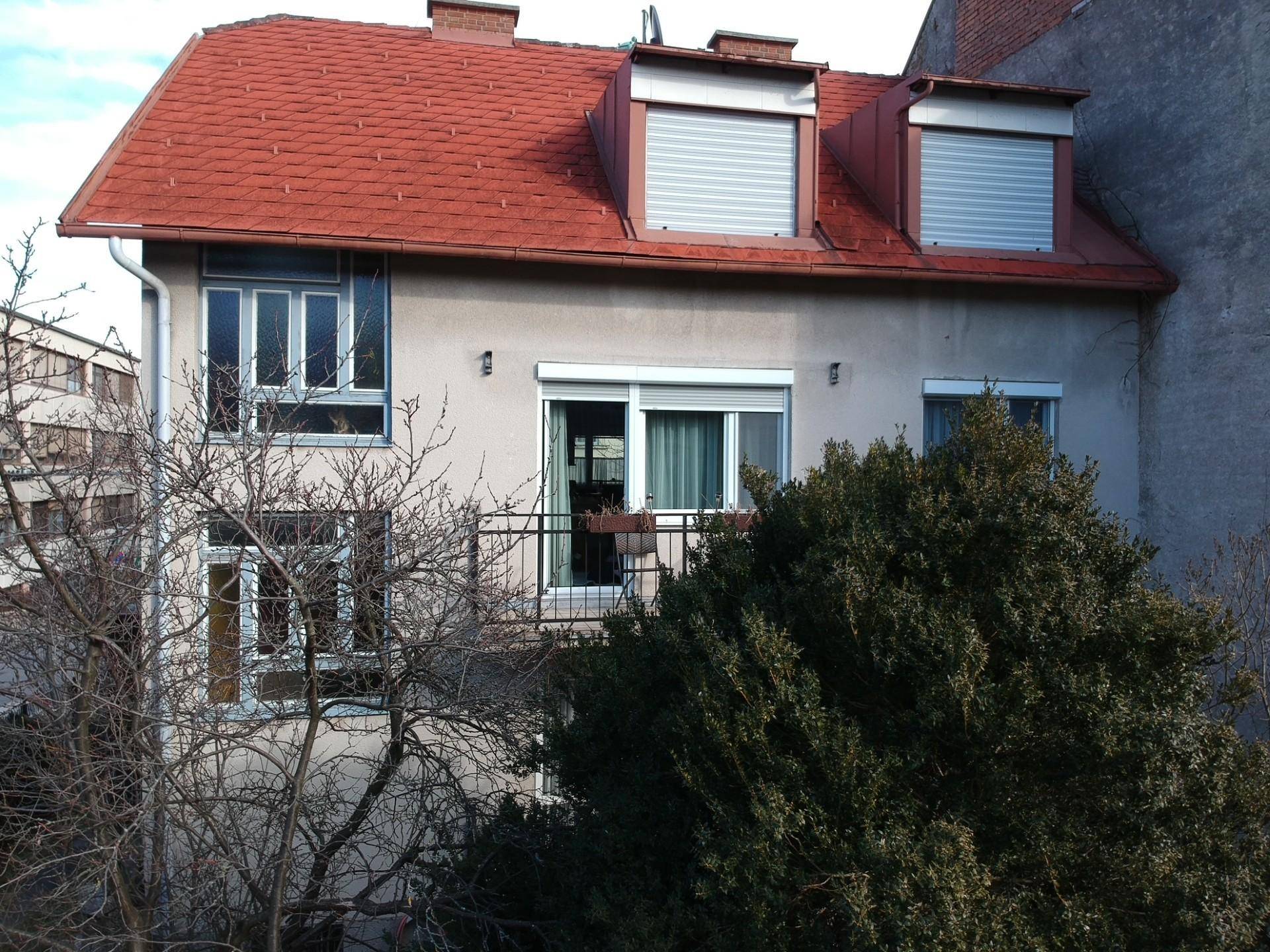 Fassade des Wohnhauses mit Balkon, roten Dachziegeln und grüner Umgebung, bei Tageslicht aufgenommen.