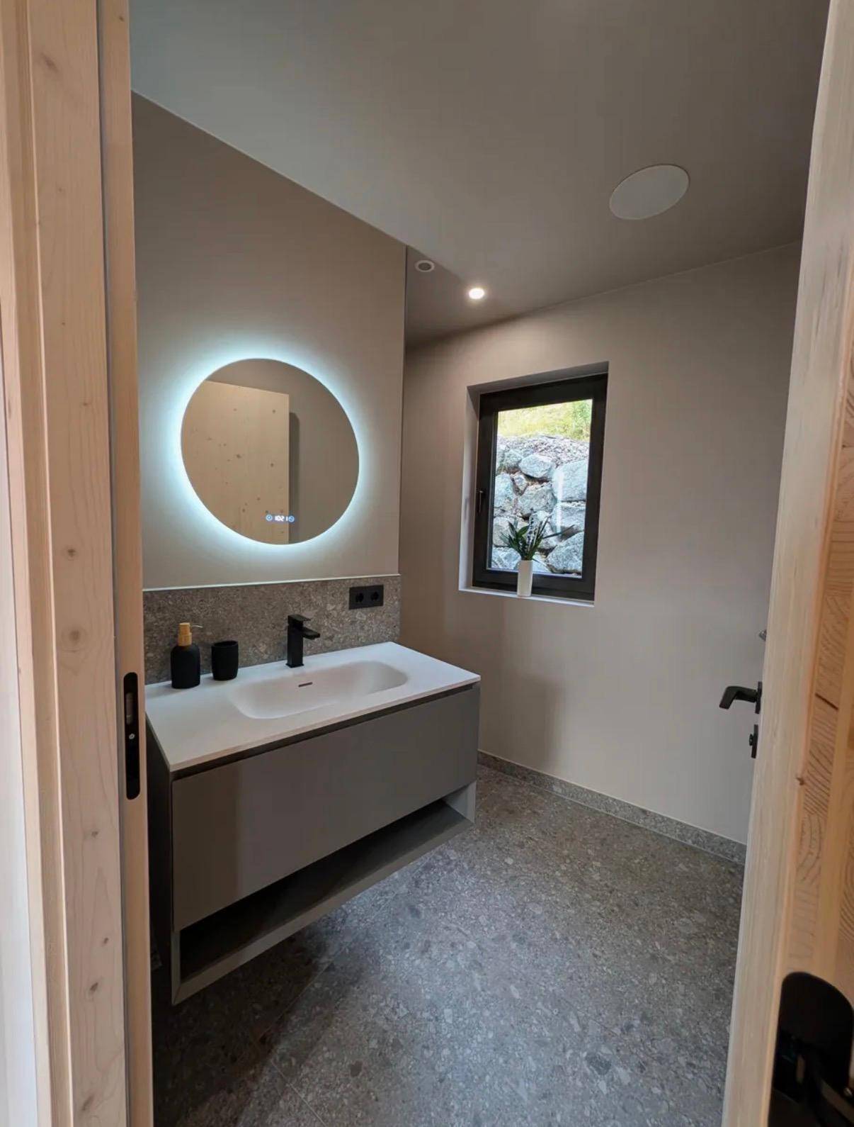 Modernes Badezimmer mit beleuchtetem Spiegel, Einzelwaschbecken und Fenster mit Blick ins Grüne.