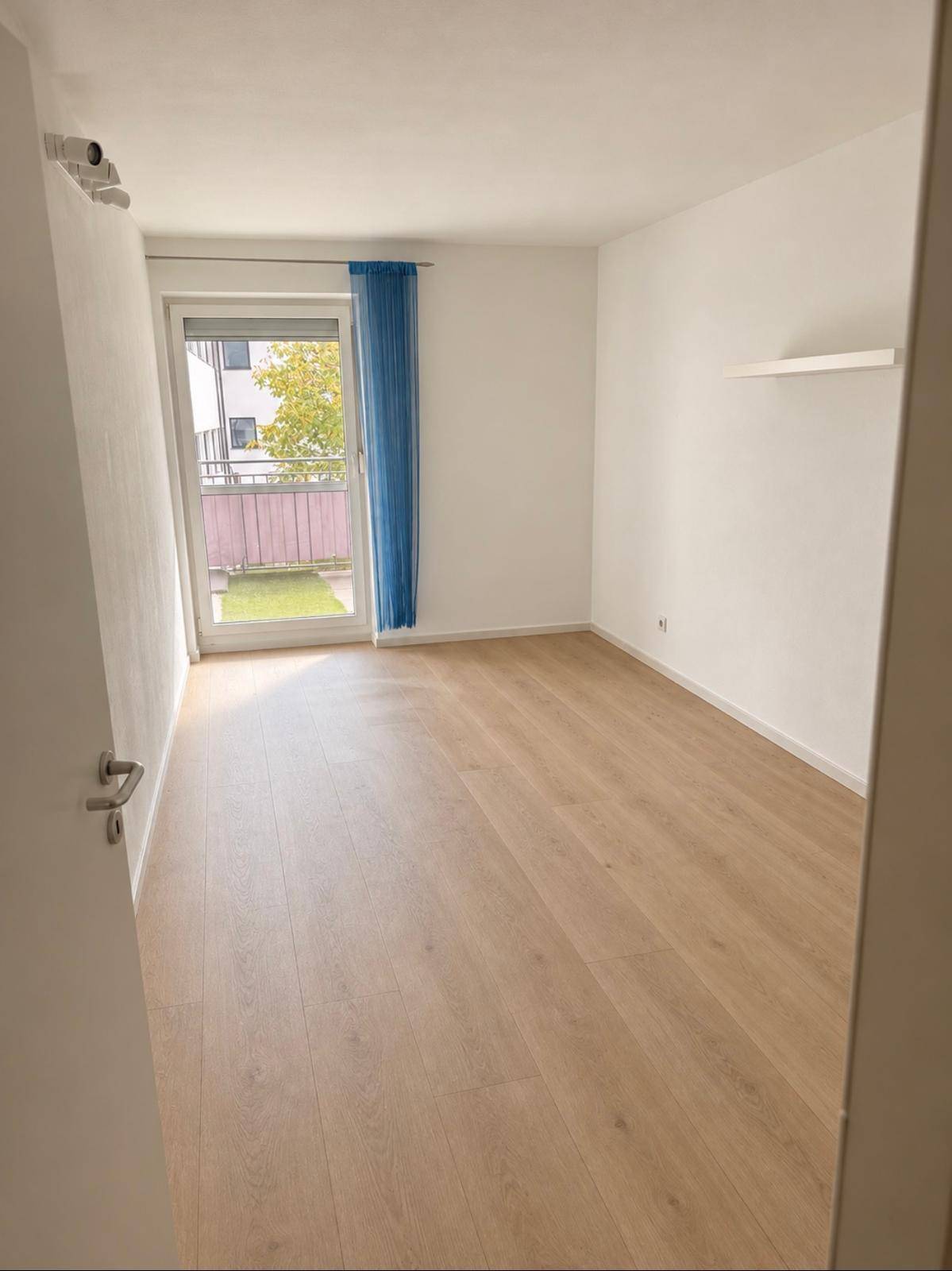 Leeres Zimmer mit hellem Laminatboden und Zugang zu einem kleinen Balkon.